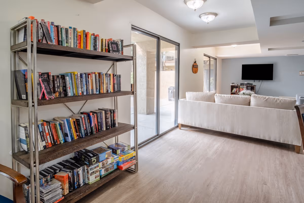 A bright common living area with a tall bookshelf stocked with books and games, sliding glass doors, and a light-colored sofa facing a wall-mounted TV.