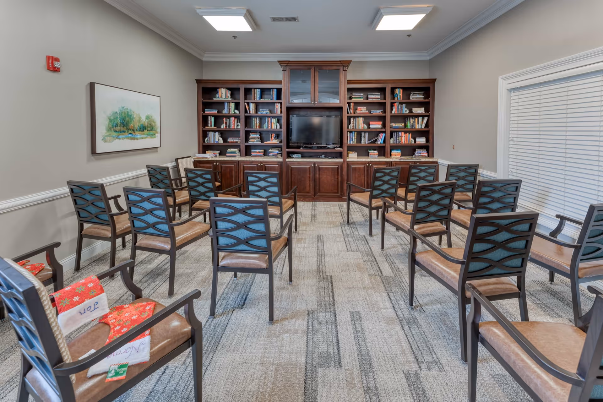 A room with multiple chairs arranged in rows facing a large wooden bookshelf with a TV in the center. The bookshelf is filled with books and the room has a carpeted floor, light gray walls, a window with blinds, and a framed painting on one wall.