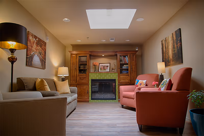 A cozy living room with a gray sofa on the left and two orange armchairs on the right, each with decorative pillows. The room features a wooden cabinet with shelves and a green-tiled fireplace in the center. There are two table lamps, a floor lamp, and two framed pictures of autumn forest scenes on the walls. A skylight is visible on the ceiling, and a potted plant is in the corner.