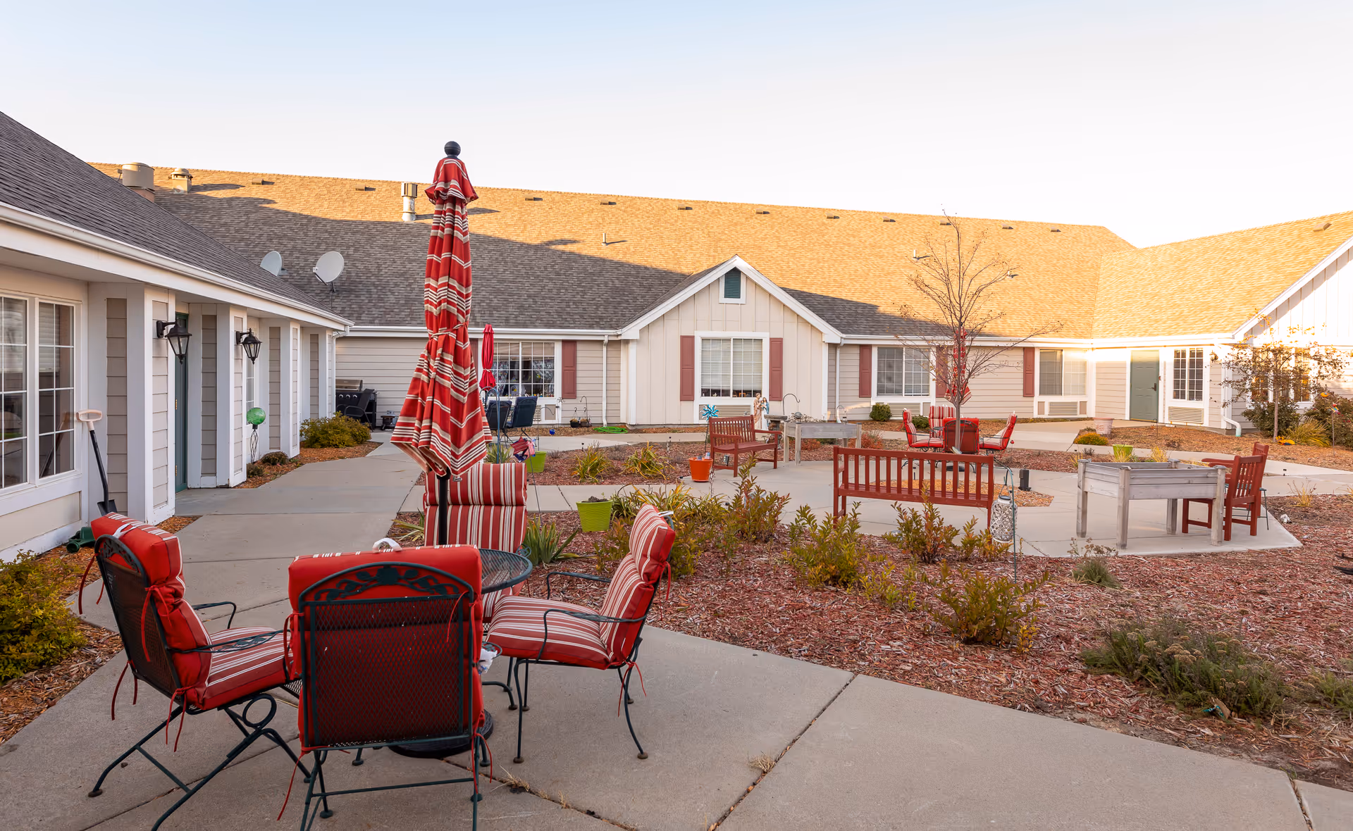 Outdoor courtyard area of an assisted living facility with red cushioned chairs around a round table with a closed red and white striped umbrella. There are benches, planters, and small shrubs in the landscaped area surrounded by a concrete walkway. The building has beige siding with white trim and red shutters.