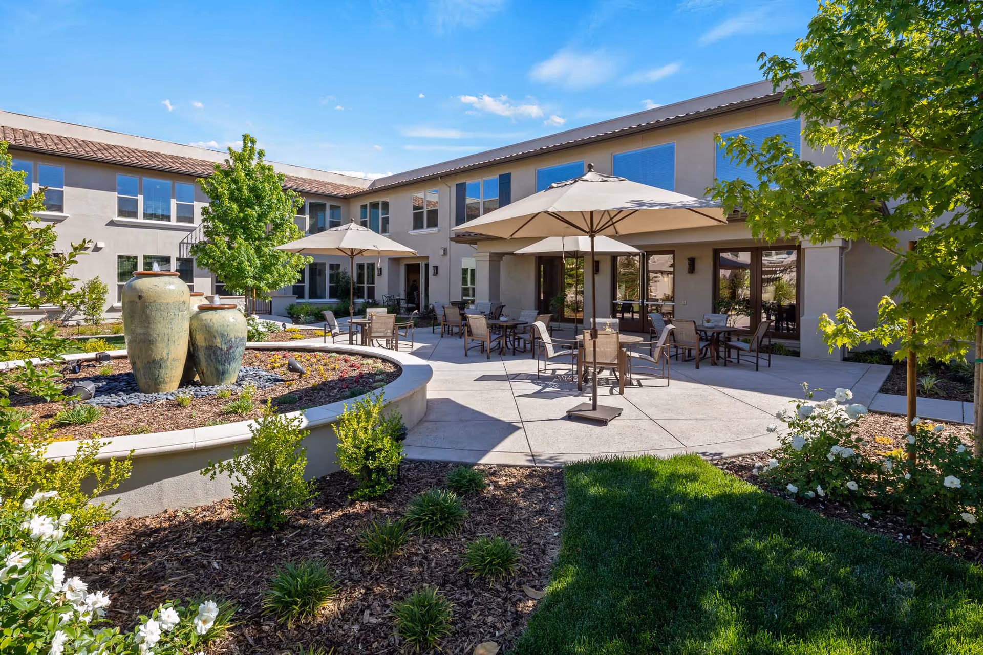 Sunny courtyard patio at Oakmont of Westpark with umbrellas and seating, a decorative urn fountain, landscaped beds, and a two-story building facade.