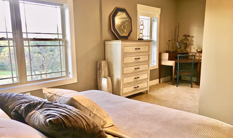 A cozy bedroom with a bed in the foreground featuring multiple pillows. There is a window on the left side letting in natural light. Against the far wall, there is a light-colored chest of drawers with a decorative mirror and vase on top. To the right, there is a small wooden desk with a chair and some decorative items, including a lamp and plants.