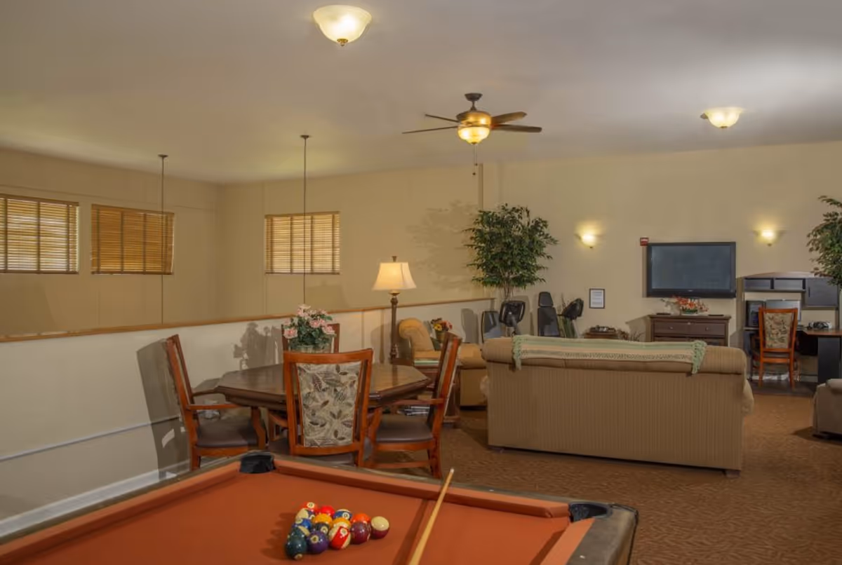 A senior living facility common area featuring a pool table with balls and a cue stick, a wooden dining table with four chairs, a beige sofa, a flat-screen TV mounted on the wall, a desk with a chair, and several plants. The room is softly lit with ceiling lights and wall sconces.