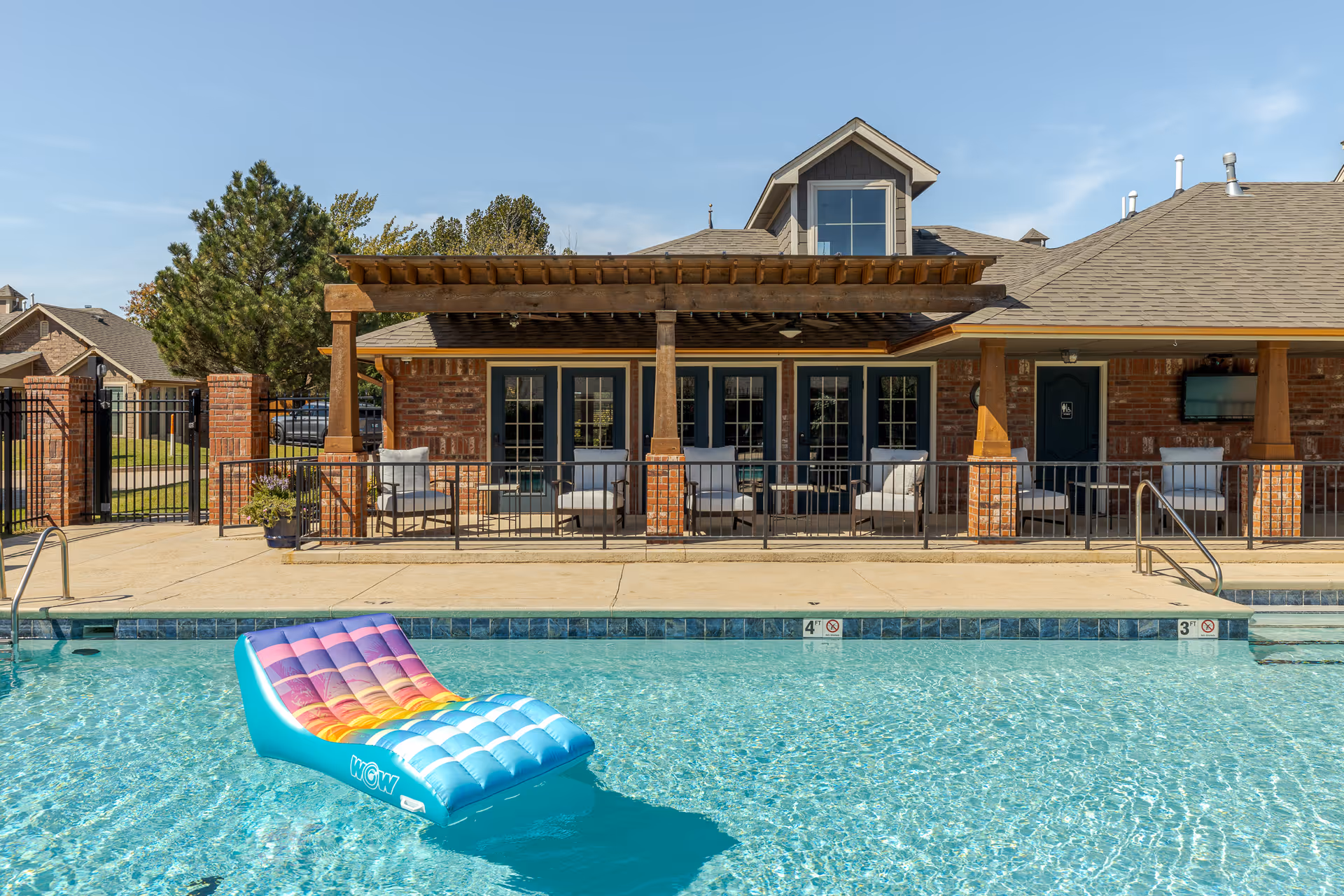 Outdoor swimming pool with a colorful inflatable lounge in front of a brick clubhouse and covered patio.