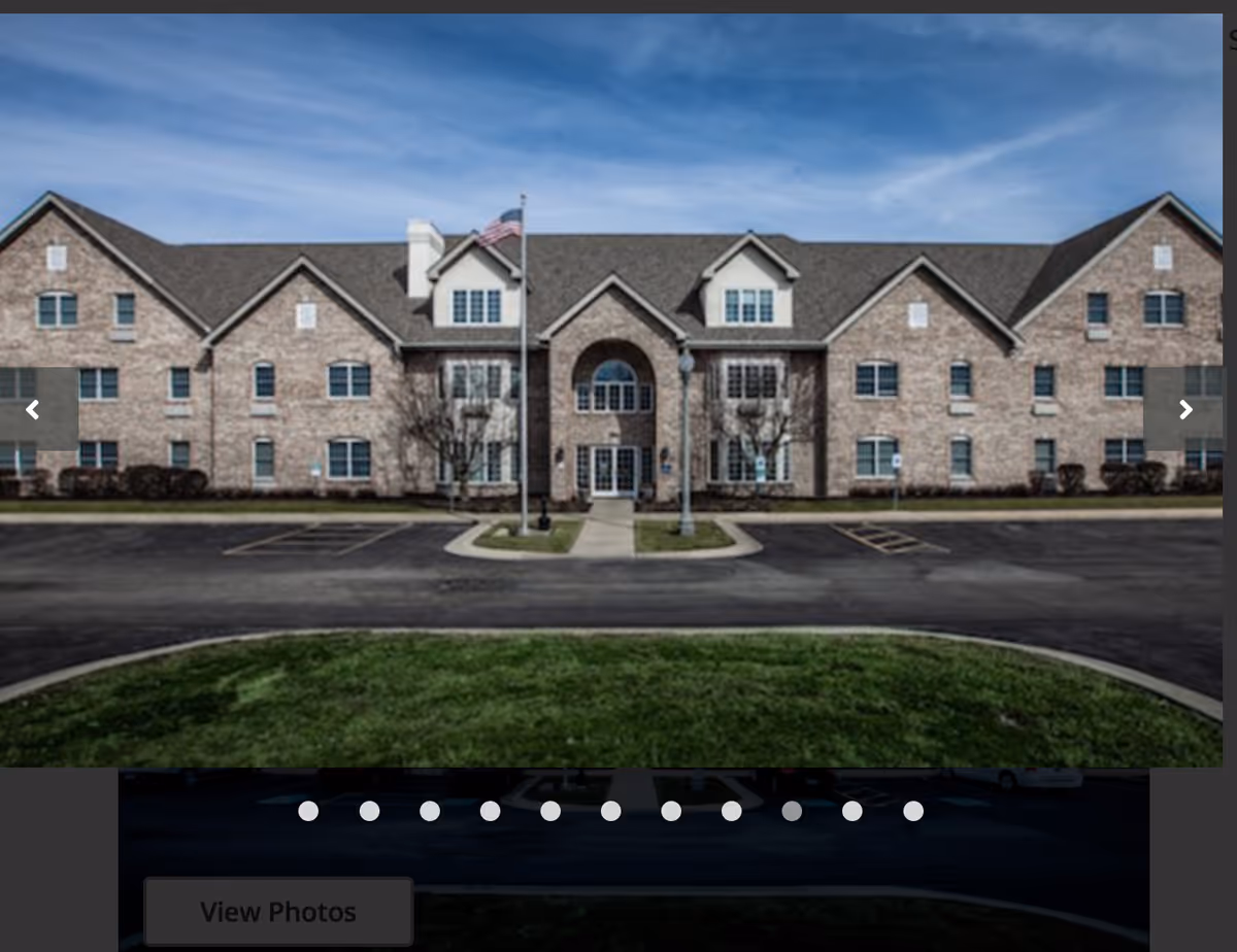 Front exterior view of a large brick senior living facility building with multiple windows, a central entrance with an archway, an American flag on a flagpole, and a parking area in front.
