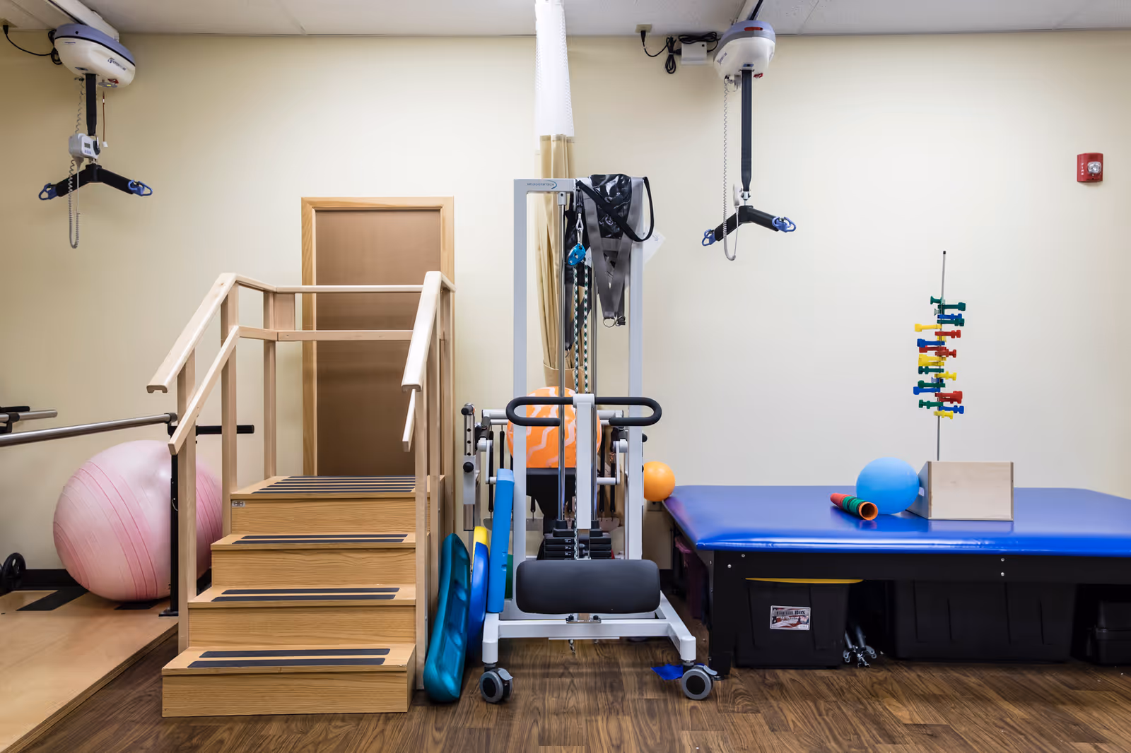 Physical therapy room with wooden stairs and handrails for rehabilitation, exercise equipment including a resistance machine, therapy balls, and a padded blue therapy table with colorful therapy tools on top. The room has light-colored walls and wood flooring.