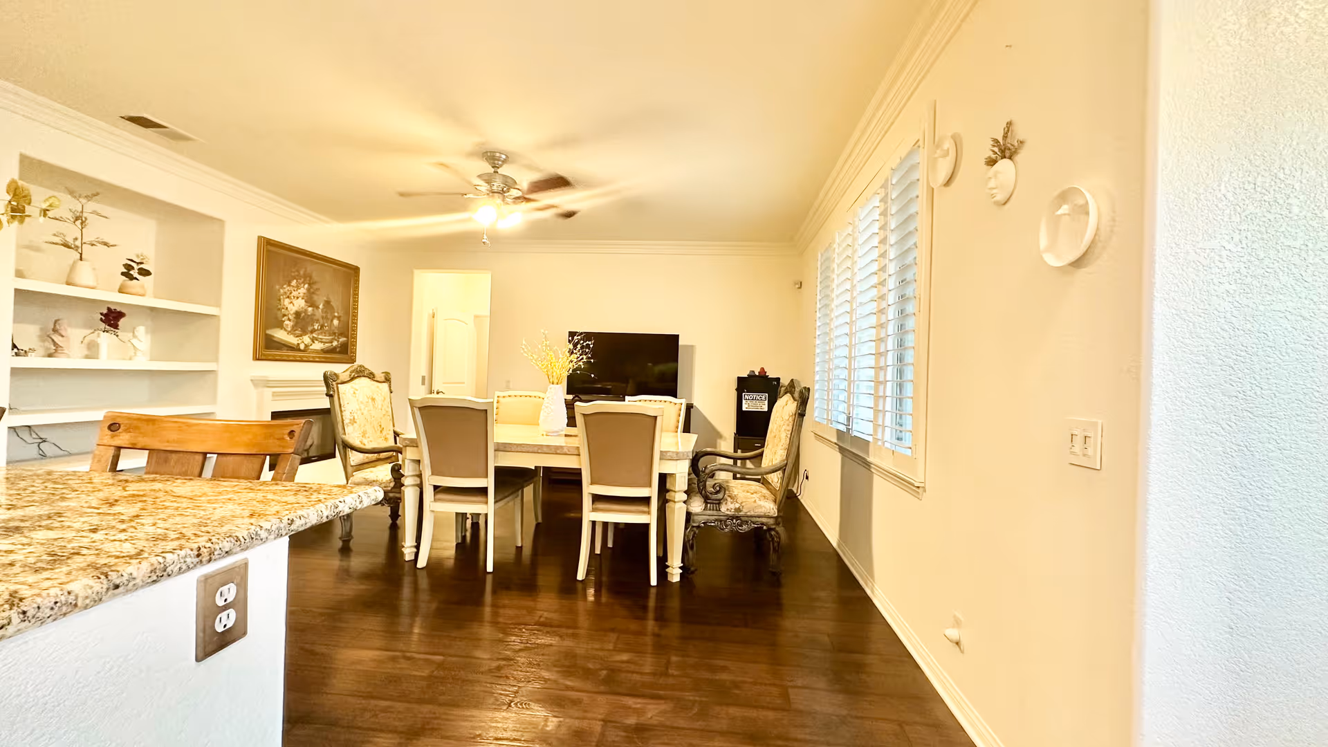 A bright dining area with a rectangular table surrounded by six chairs, two of which have ornate armrests. The room features dark wooden flooring, a ceiling fan with a light, and a large window with white shutters on the right wall. There is a granite countertop with a wooden chair in the foreground on the left, built-in white shelves with decorative items on the far left, and a flat-screen TV against the back wall.