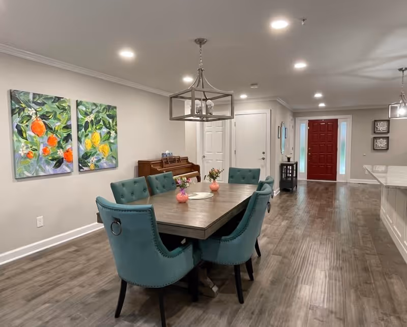 A dining area with a rectangular wooden table surrounded by six teal upholstered chairs. On the table are three small pink vases with flowers. The room has wood flooring and light gray walls. Two colorful paintings of fruit hang on the left wall above a piano. In the background is a red front door with sidelights, and a kitchen counter is visible on the right side.