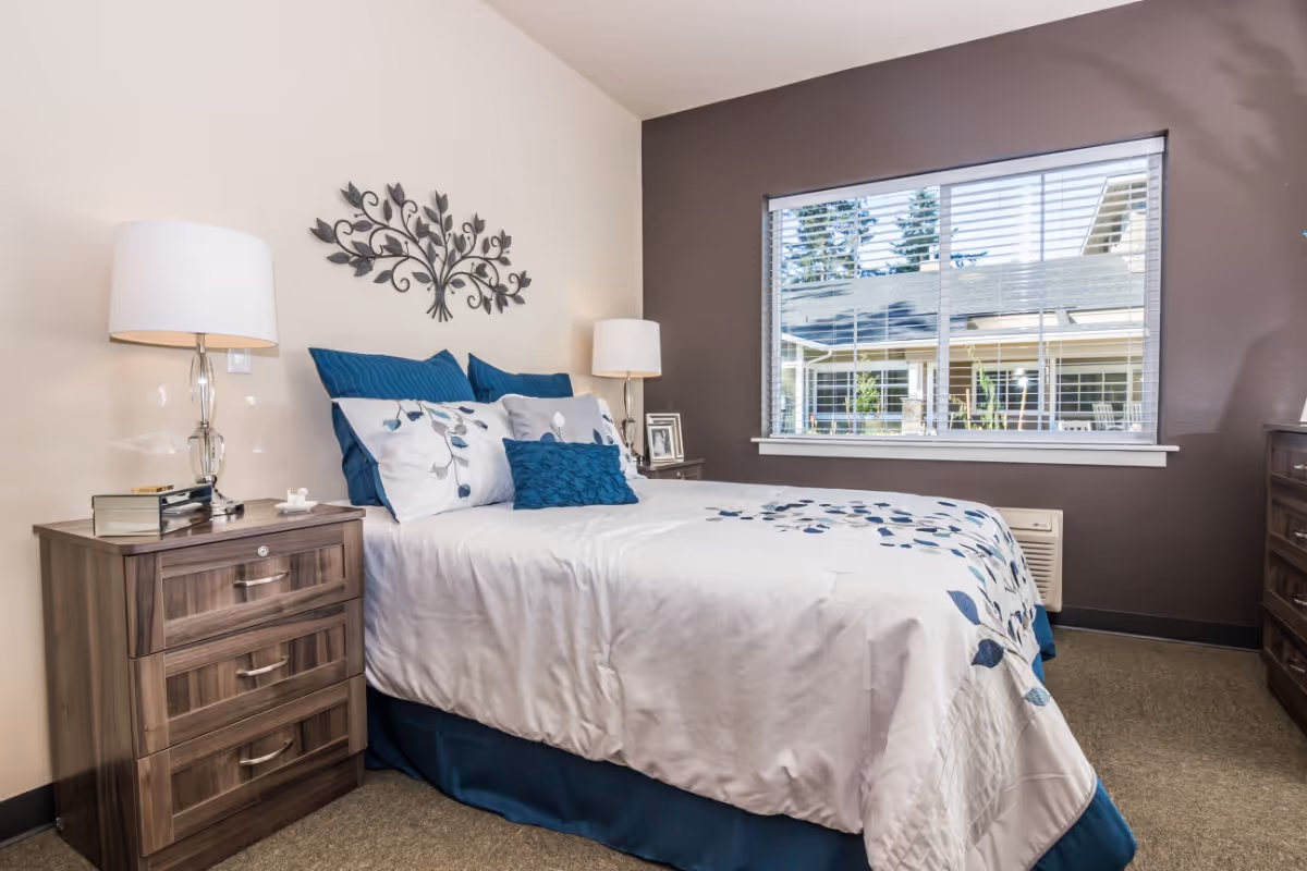 A neatly made bed with white and blue floral bedding in a bedroom. There are two wooden nightstands on either side of the bed, each with a lamp. A decorative metal wall art piece is mounted above the bed. A large window with white blinds shows an outdoor view of another building and trees.