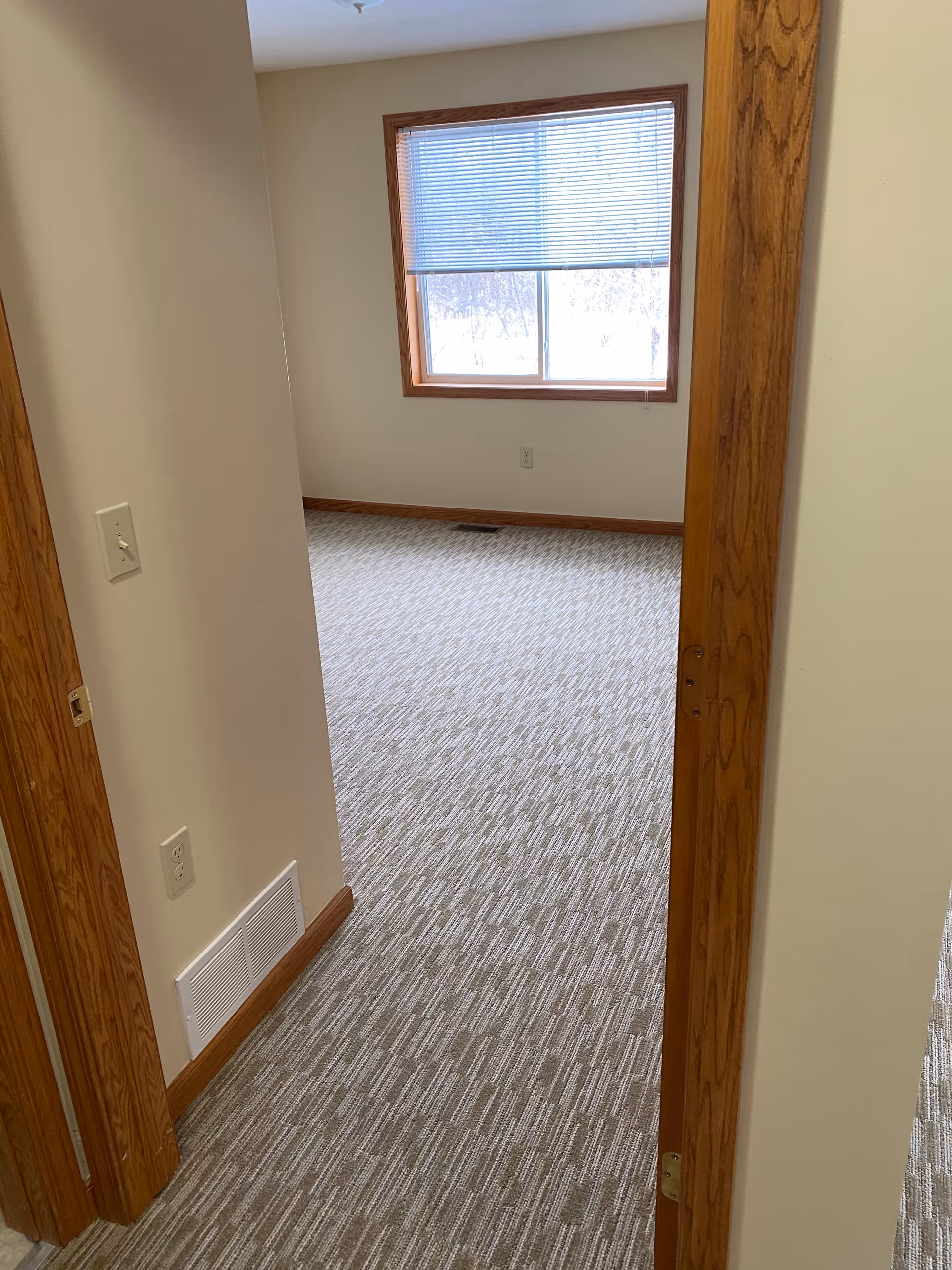 View through a doorway into an empty room with beige walls, a window with partially closed blinds, and a carpeted floor with a textured pattern. The door frame and baseboards are wooden with a natural finish.