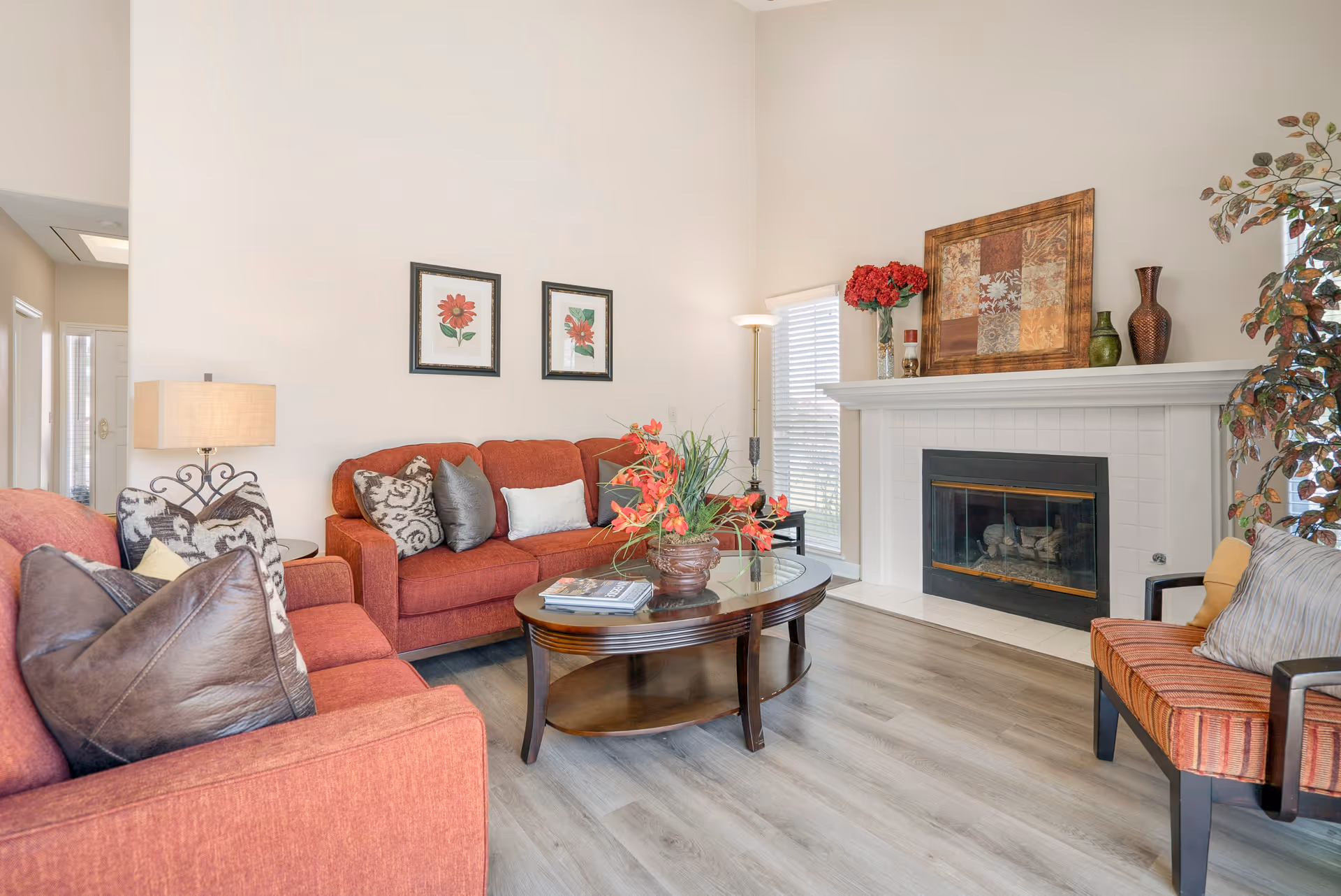 A bright living room with two red sofas, a wooden coffee table with a flower arrangement, a fireplace with decorative vases and artwork above it, a floor lamp, and a window with blinds. The room has light-colored walls and wood flooring.