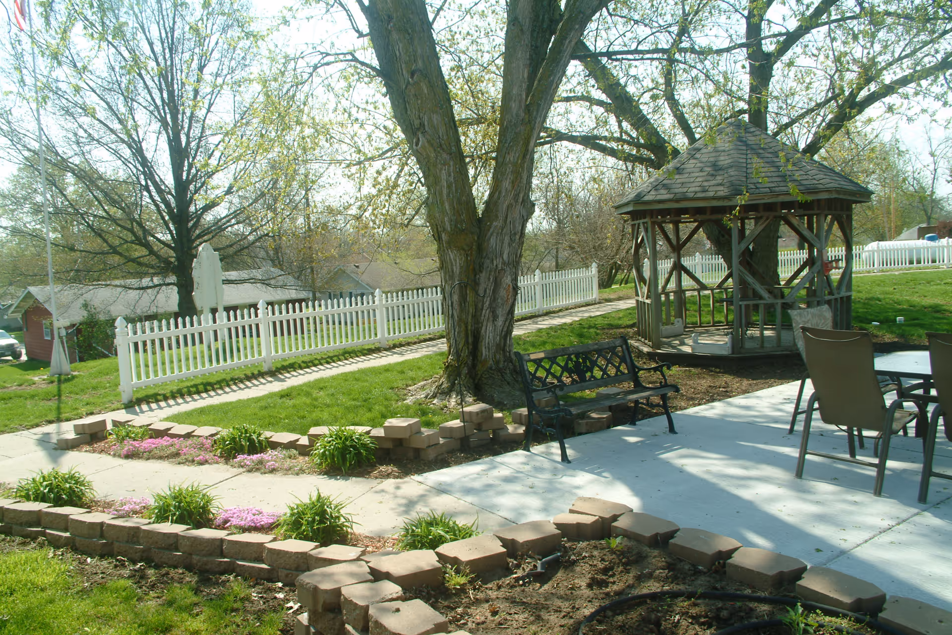 Sunny outdoor courtyard with a large tree, wooden gazebo, metal bench, patio chairs, and a white picket fence.
