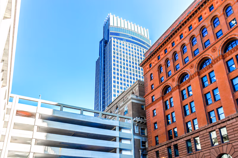 View of several buildings in a city under a clear blue sky, including a tall modern skyscraper with a curved top, a red brick building with arched windows, and a white multi-level parking structure.