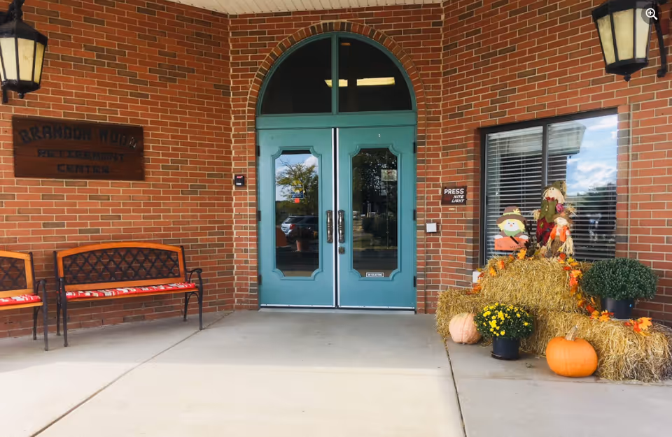 Entrance to Brandon Wood Retirement Center with teal double doors, brick facade, benches and fall decorations including hay bales and pumpkins.