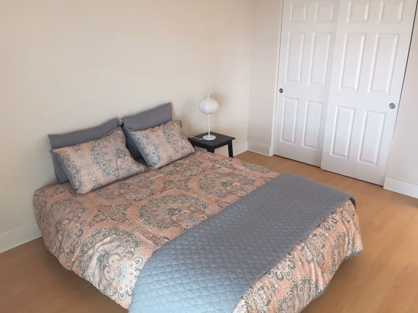 A bedroom with a bed covered in a patterned pink and gray bedspread with matching pillows and a gray quilted throw at the foot. Next to the bed is a small black nightstand with a white spherical lamp. The room has light-colored walls, a wooden floor, and white double sliding closet doors.
