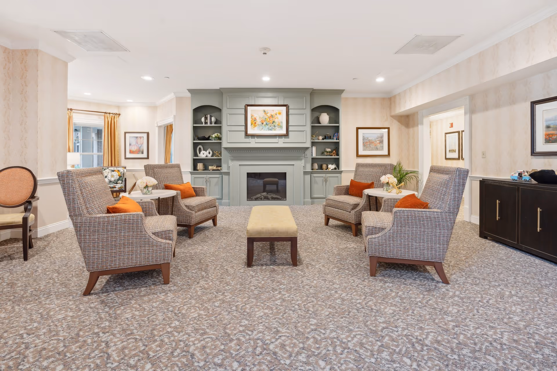 A cozy living room area with four upholstered armchairs arranged around two small round tables, each table decorated with a small flower arrangement. The chairs have orange accent pillows. In the background, there is a green built-in cabinet with shelves and a fireplace in the center. Above the fireplace hangs a colorful floral painting. The room has patterned carpet flooring, beige wallpaper, and framed artwork on the walls.
