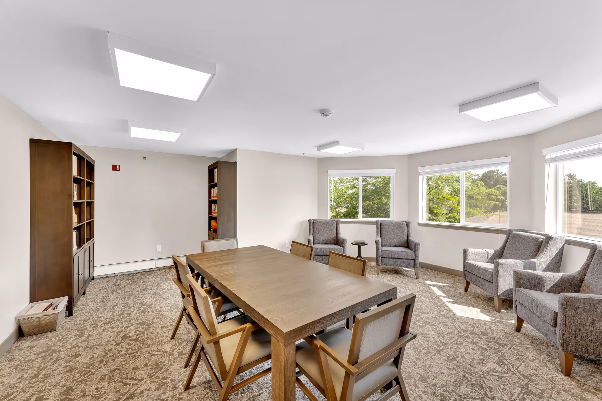A bright and spacious common room with a large wooden table surrounded by eight chairs in the center. There are four upholstered armchairs arranged near three large windows that let in natural light. Two tall wooden bookshelves filled with books are against the wall. The room has beige walls and carpeted flooring with modern ceiling lights.