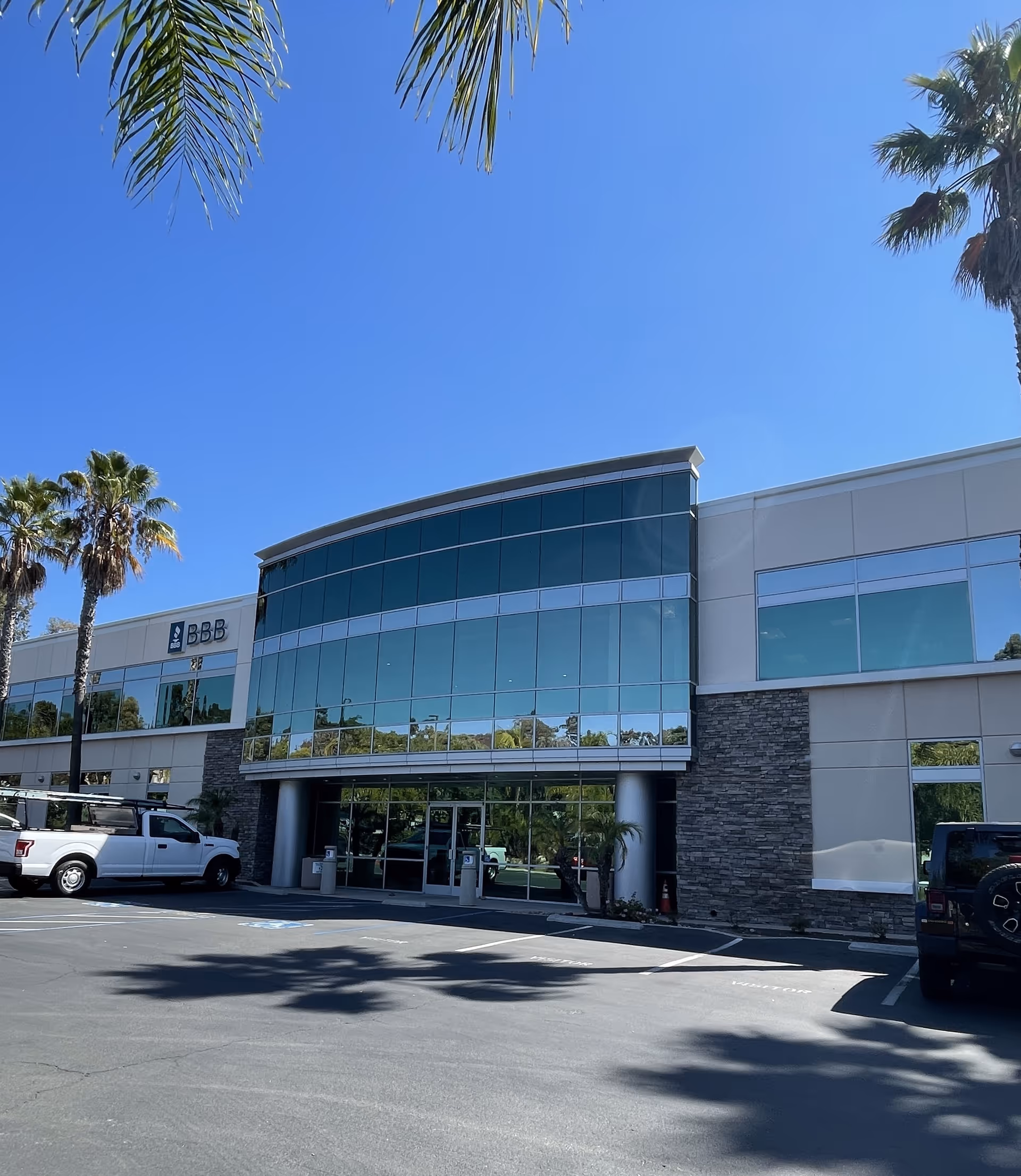 Modern two-story commercial building with large glass windows and stone accents, surrounded by palm trees under a clear blue sky. Several vehicles are parked in front of the building.