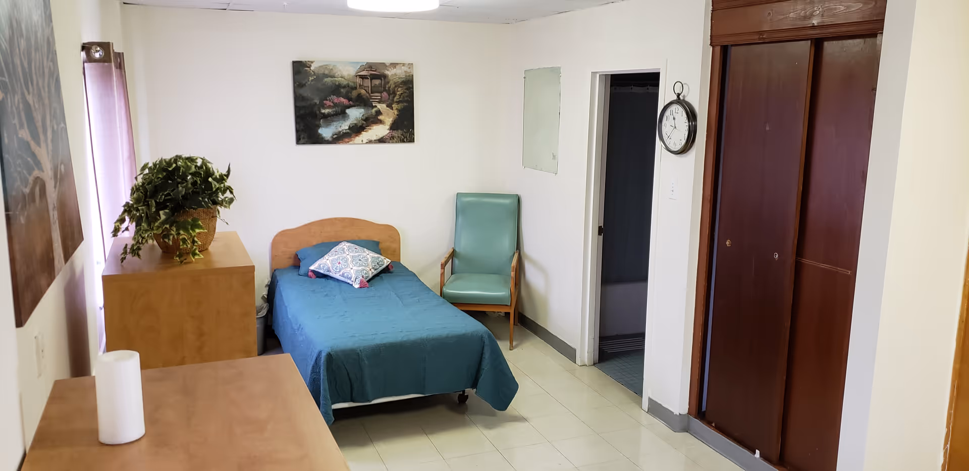 A small bedroom with a single bed covered in a blue bedspread and a decorative pillow. Next to the bed is a green armchair and a wooden dresser with a potted plant on top. A painting of a garden scene hangs on the wall above the bed. There is a clock on the wall near a doorway leading to a bathroom with a shower curtain visible. The room has tiled flooring and wooden closet doors.
