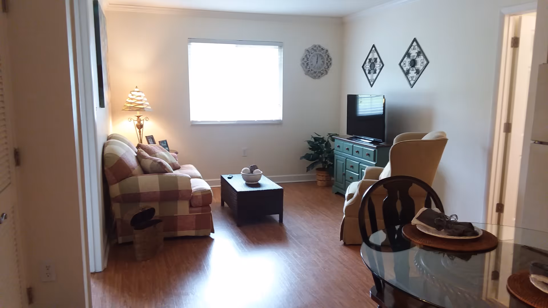 A cozy living room with a plaid sofa, a floor lamp, a coffee table with decorative items, a green TV stand with a flat-screen television, an armchair, and a round glass dining table with chairs. The room has wooden flooring, a window with blinds, and decorative wall hangings including a clock and two diamond-shaped pieces.