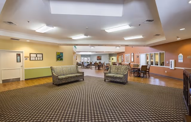 Spacious interior common area with patterned carpet and two upholstered sofas facing each other. In the background, there are multiple tables and chairs arranged in a dining or activity space. The walls are painted in warm tones with artwork and bulletin boards displayed. The ceiling has recessed lighting and a skylight.