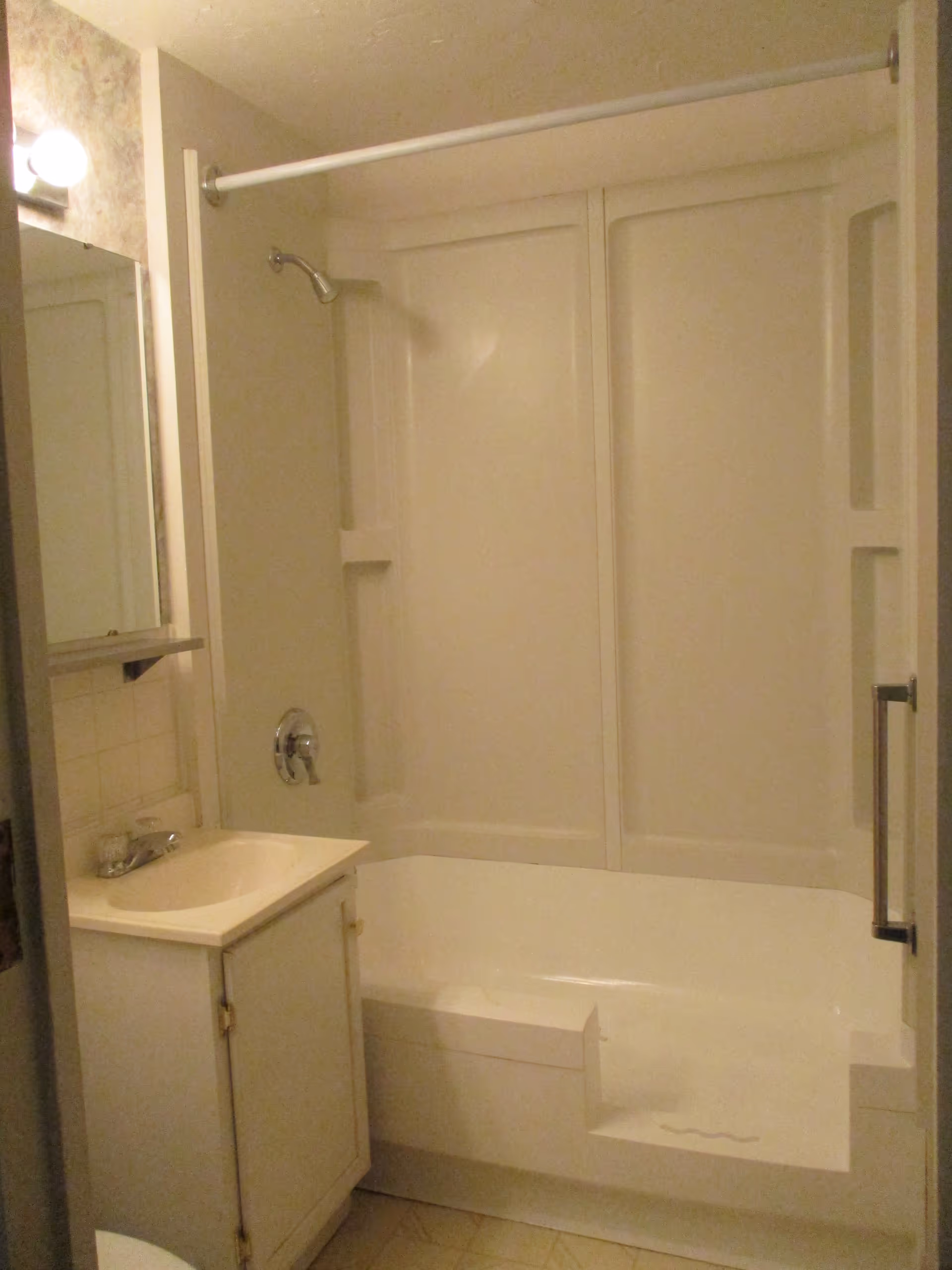 A small bathroom featuring a white bathtub with a built-in seat and a showerhead above. There is a white vanity with a sink and a mirror above it on the left side. The walls are light-colored, and a light fixture with a round bulb is visible above the mirror.