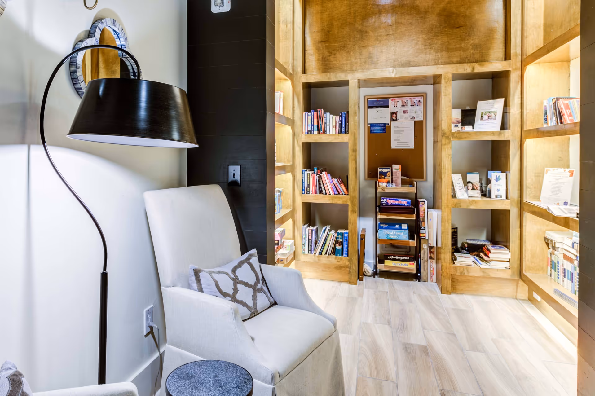 Cozy reading nook with an upholstered armchair and floor lamp facing built-in wooden bookshelves filled with books and brochures.
