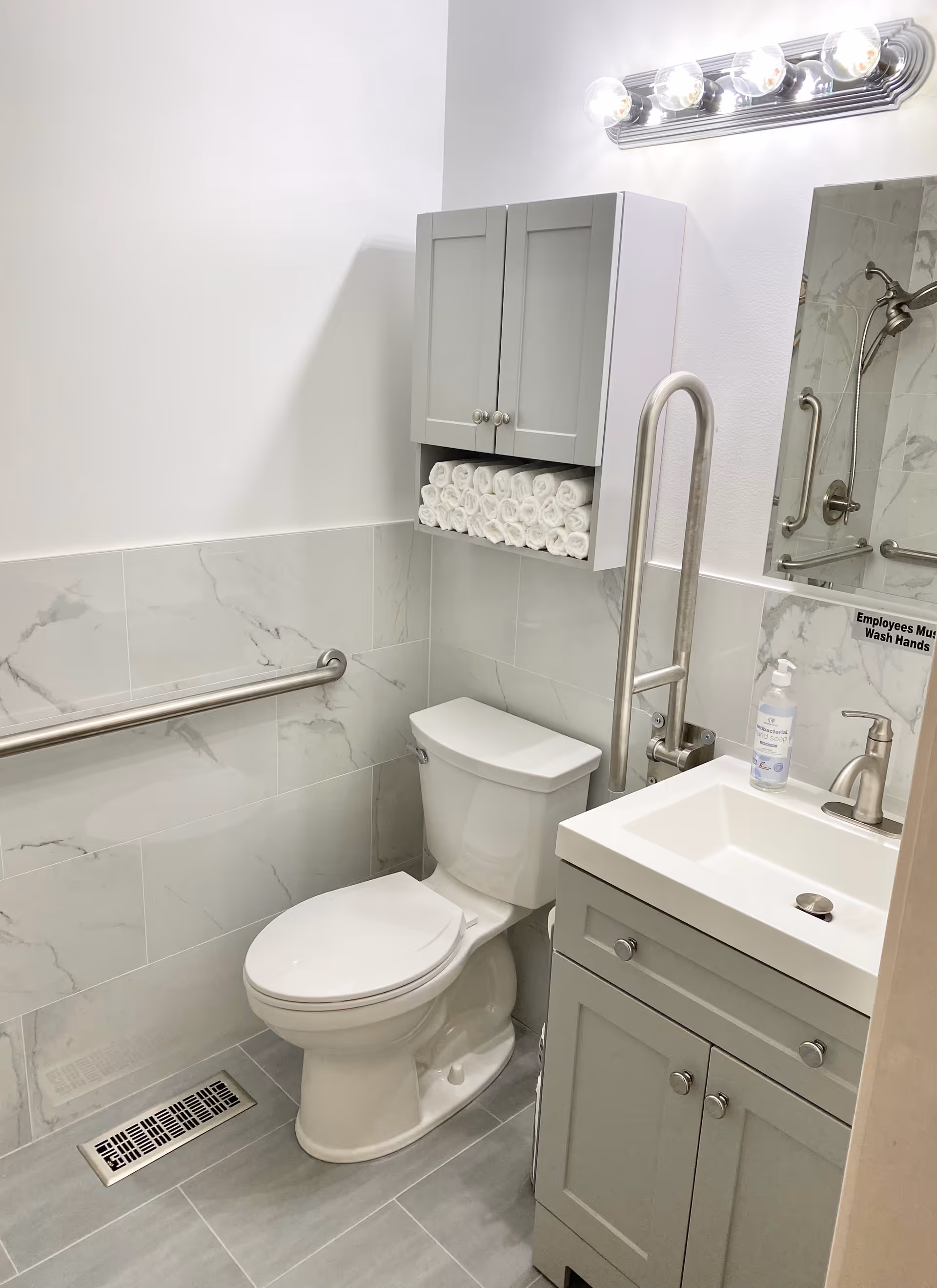 Accessible bathroom with a toilet, stainless grab bars, a sink vanity, wall cabinet stocked with rolled towels, mirror and light fixture.