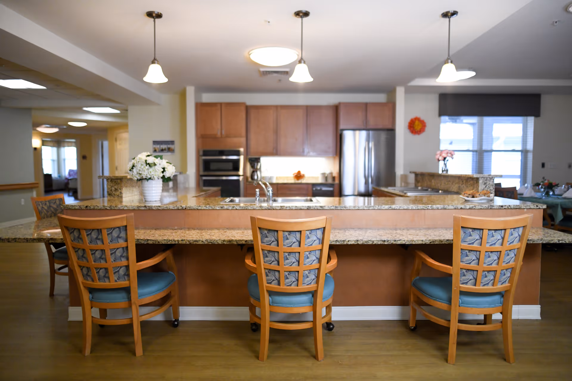 A spacious kitchen area with a granite countertop island and three wooden chairs with blue cushioned seats. The kitchen features wooden cabinets, a stainless steel refrigerator, built-in oven, and a coffee maker. Pendant lights hang from the ceiling, and there are flowers in vases on the counter. In the background, there is a dining area with tables and chairs near windows.