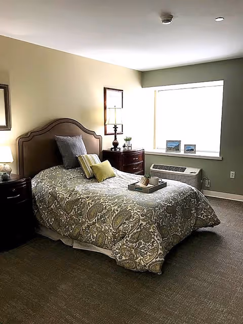 A neatly made single bed with a patterned bedspread and several pillows in a senior living bedroom. There are two wooden nightstands on either side of the bed, each with a lamp. A window with framed pictures on the sill and an air conditioning unit below it is visible on the right wall.