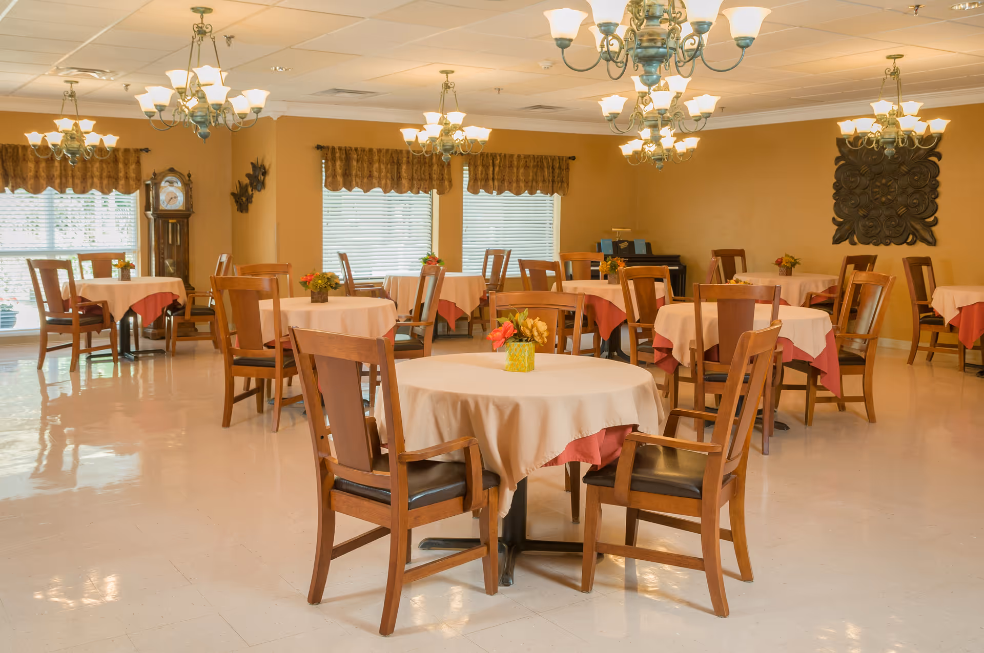 Bright dining room with round tables covered in tablecloths, wooden chairs, chandeliers, and small floral centerpieces.