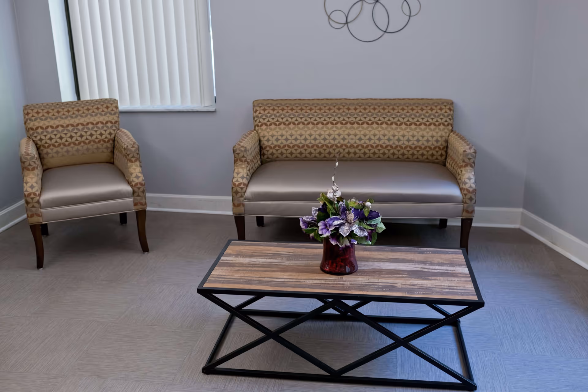 A small seating area with a patterned armchair and a matching loveseat against a light gray wall. In front of the seating is a rectangular wooden coffee table with a black metal frame, holding a vase with purple and white flowers. A window with vertical blinds is partially visible on the left side, and a decorative metal wall art piece with circular shapes hangs above the loveseat.