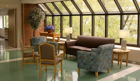 A bright sitting area with large windows letting in natural light. The room features a brown sofa, two patterned armchairs, and two wooden chairs arranged around a small table. There are two side tables with lamps and a wooden table with a potted plant and flowers near a brick wall.