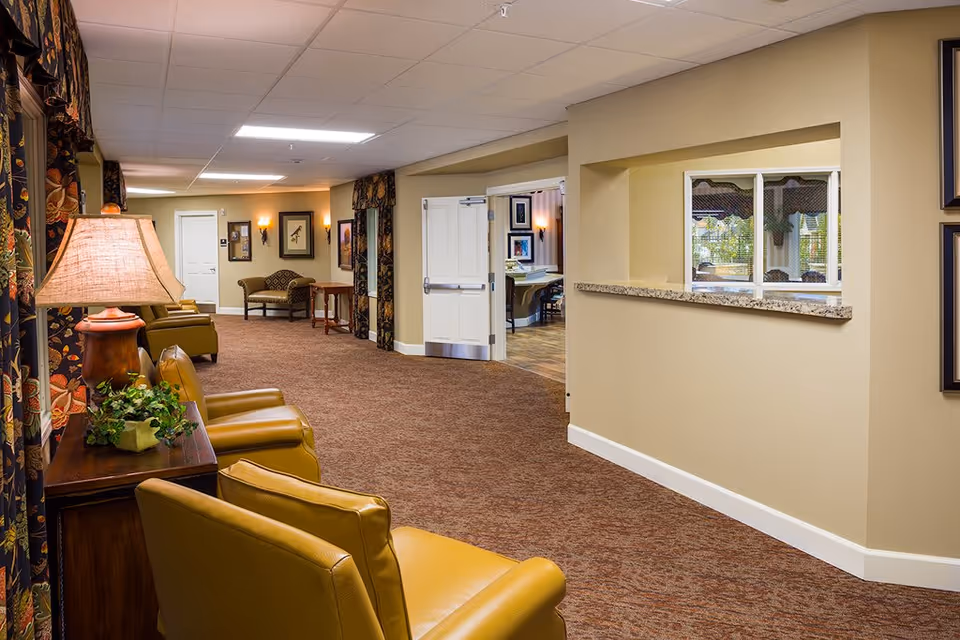 Interior hallway of a senior living facility with beige walls and brown carpet. The hallway features several mustard-yellow armchairs along the left side, a wooden side table with a lamp and a small plant, and framed artwork on the walls. There is a window opening on the right wall with a granite ledge, and double doors leading to another room in the background.