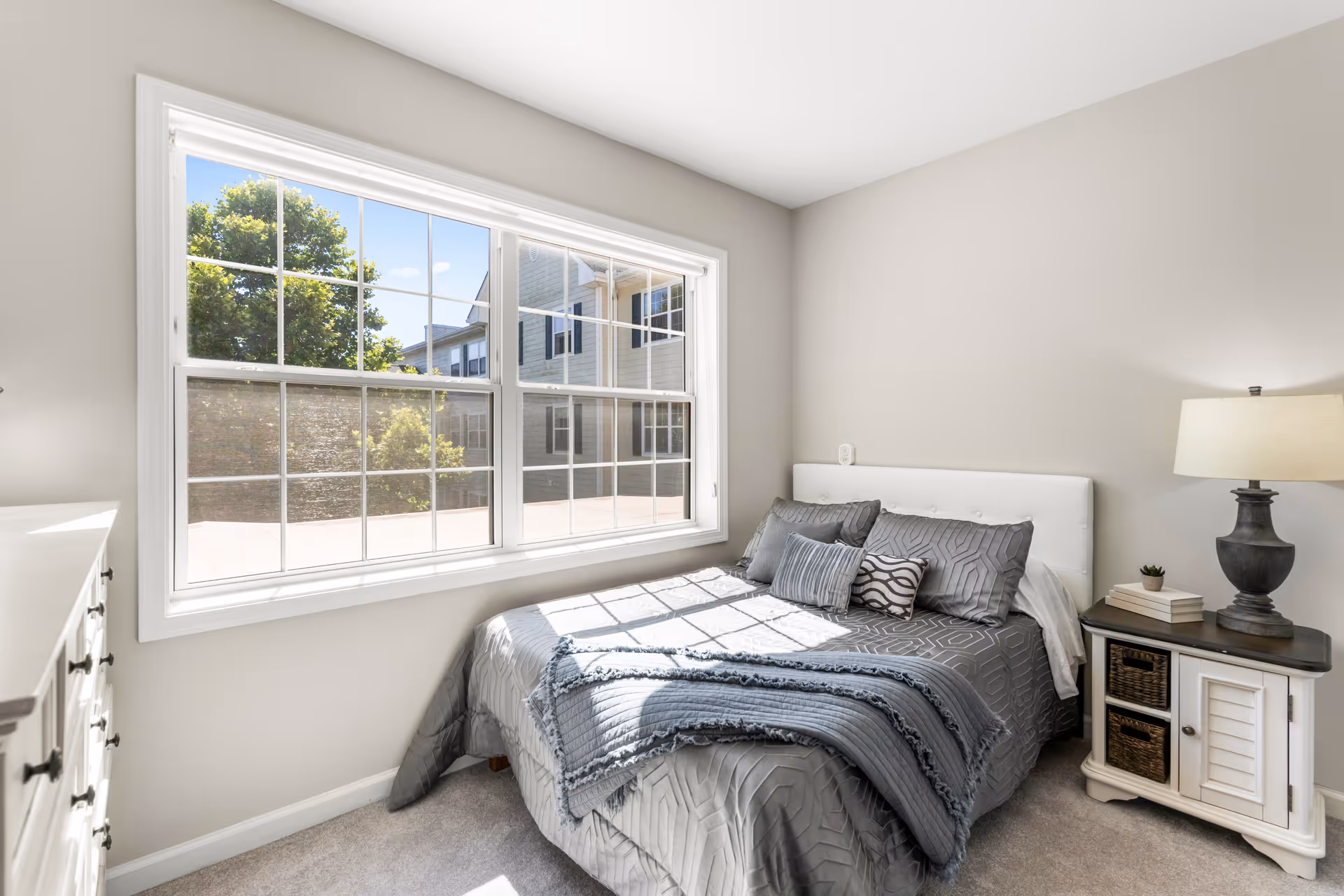 A bright bedroom with a large window letting in natural light. The bed is made with gray patterned bedding and several pillows. Next to the bed is a white nightstand with a dark lamp, a small plant, and some books. A white dresser is partially visible on the left side of the image.