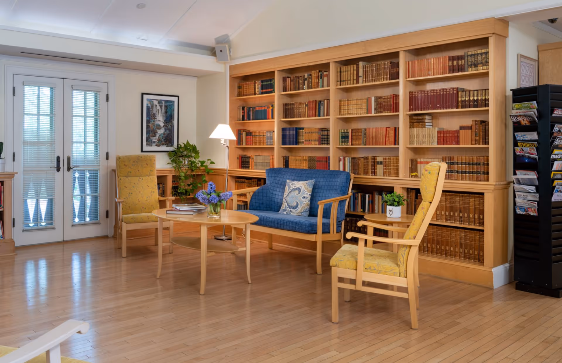 Sunlit lounge with wooden bookshelves, a blue loveseat, yellow armchairs, and a round coffee table in front of French doors.