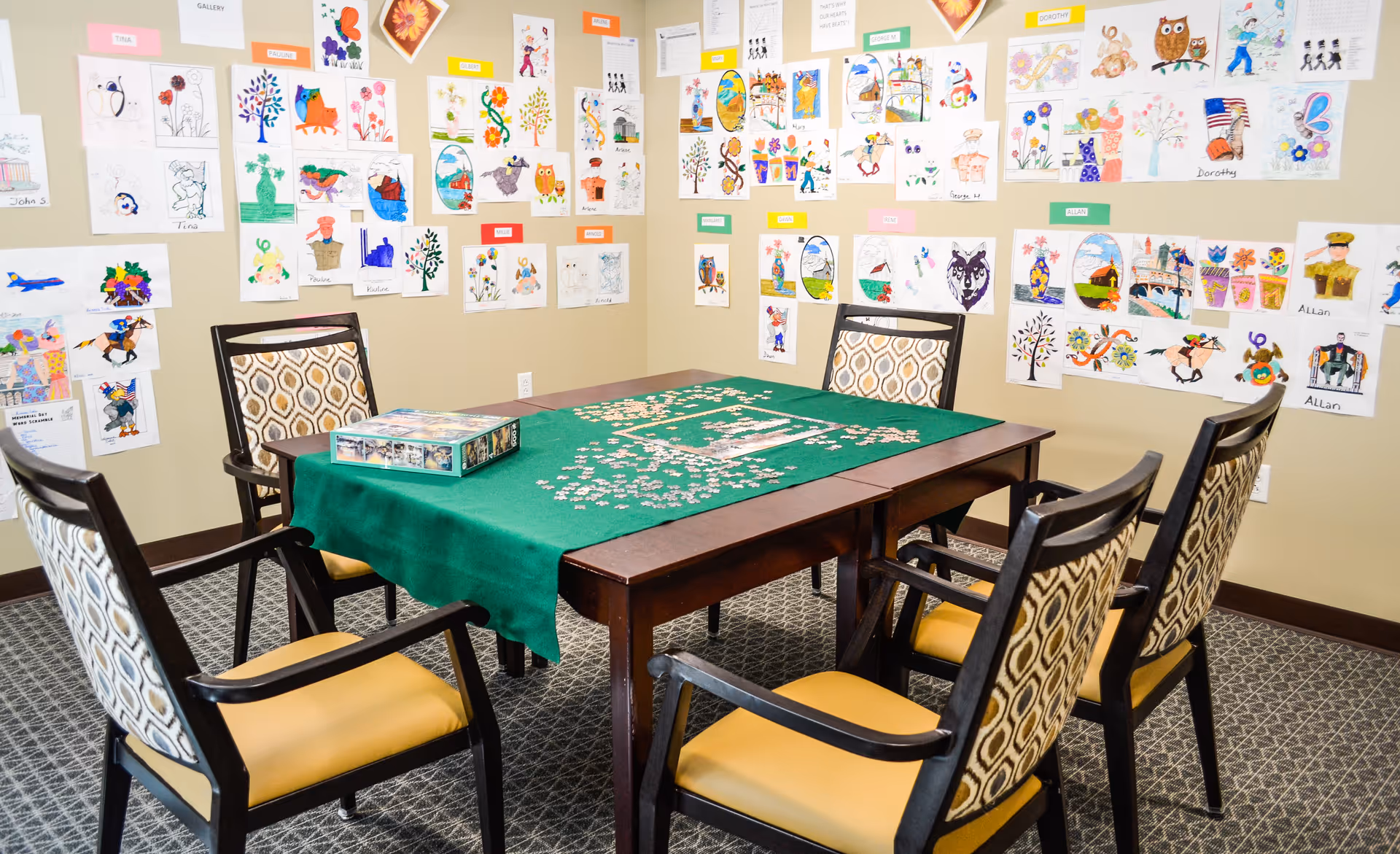A room with a wooden table covered by a green cloth and a partially completed jigsaw puzzle on top. Surrounding the table are six chairs with patterned backs and yellow seats. The walls are decorated with numerous colorful drawings and artwork, each labeled with different names.