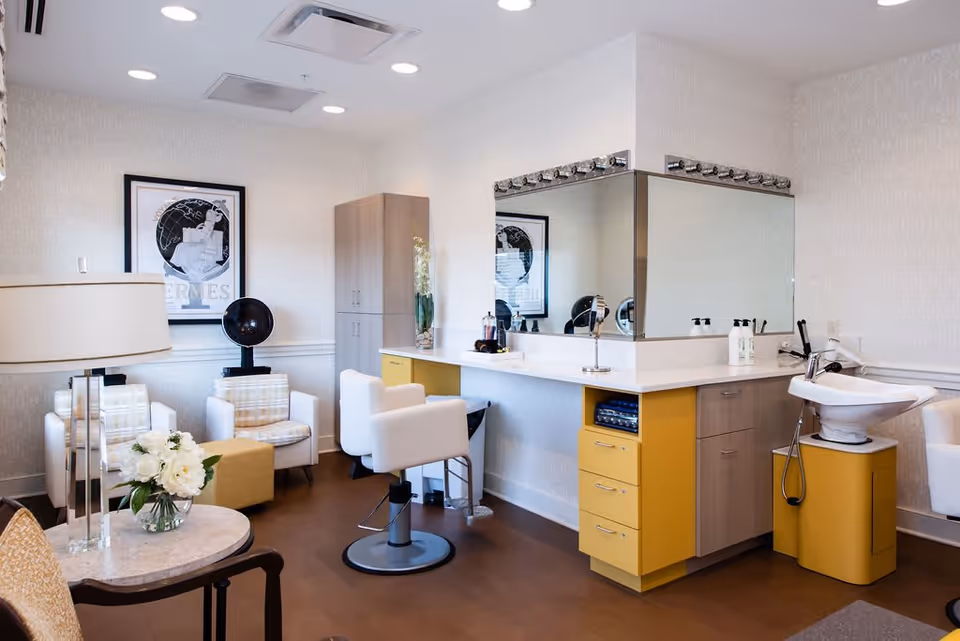 A bright and modern salon area in a senior living facility featuring a styling chair in front of a large mirror with lights, a hair washing station with a sink, two white armchairs with plaid cushions, a small ottoman, a tall cabinet, and a round table with a lamp and a vase of white flowers.