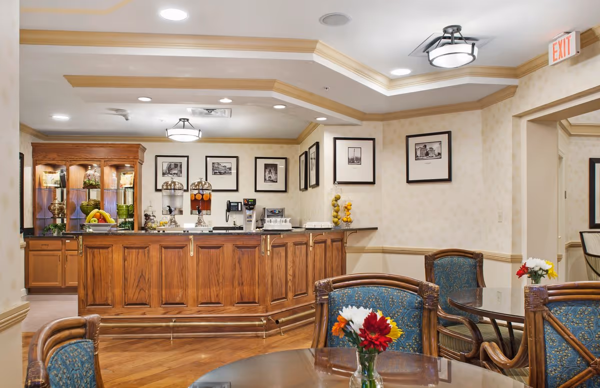 A cozy dining area in a senior living facility with wooden tables and chairs upholstered in blue patterned fabric. The room features a wooden counter with beverage dispensers and a display cabinet with decorative items. Framed black and white photographs hang on the cream-colored walls, and small vases with colorful flowers are placed on the tables. The ceiling has recessed lighting and a decorative light fixture, with an exit sign visible above a doorway.