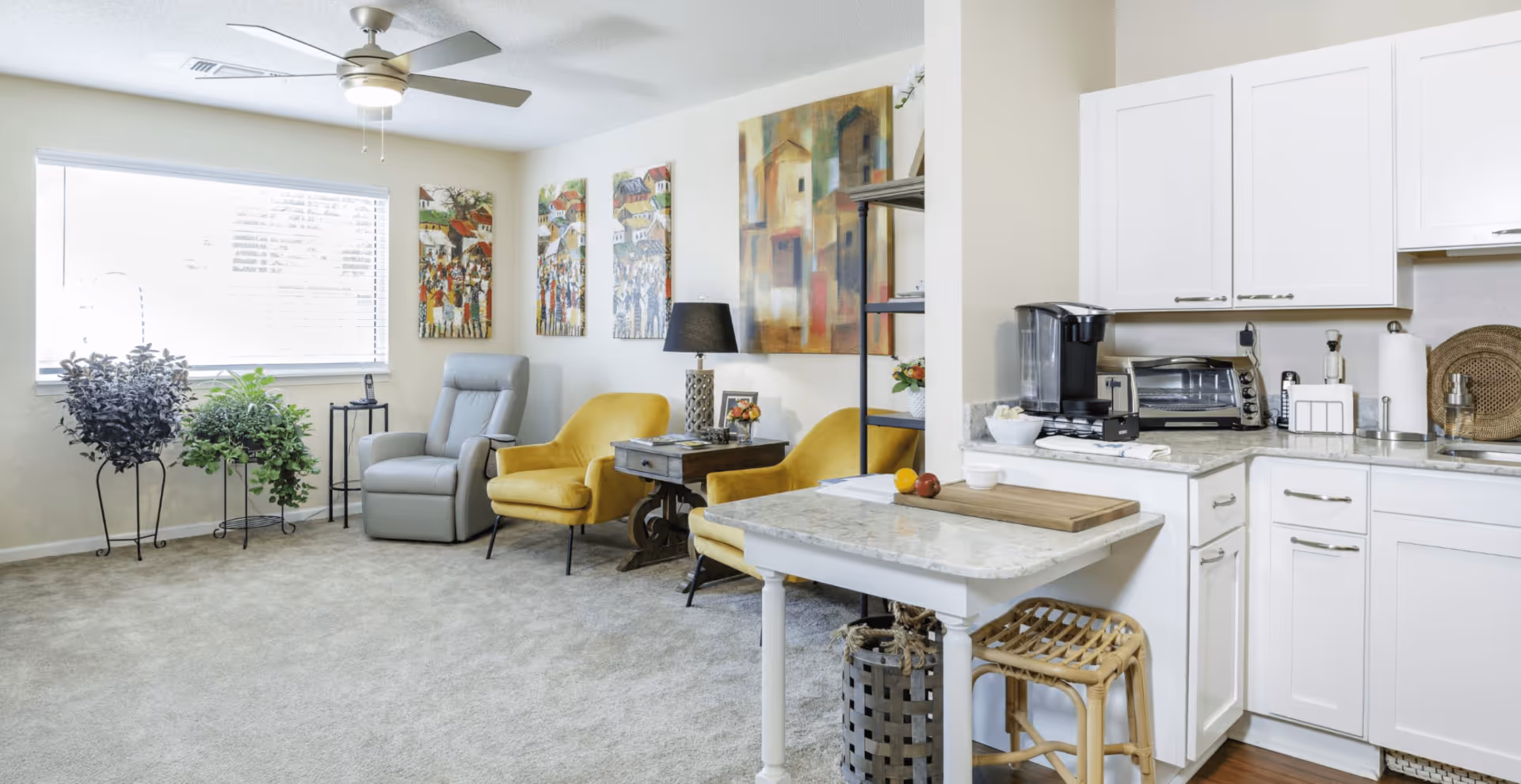 A bright and cozy living area with a large window letting in natural light. The room features a gray recliner chair, two yellow armchairs, a small side table with a lamp, and colorful artwork on the walls. Adjacent to the living area is a compact kitchen with white cabinets, a coffee maker, toaster oven, and a small island with a cutting board and a stool underneath. There are also two potted plants near the window.