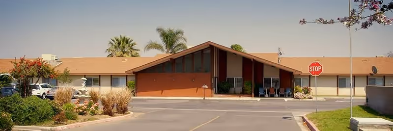 Exterior view of a single-story assisted living facility building with a triangular roof entrance, surrounded by palm trees, bushes, and a parking lot with a stop sign.