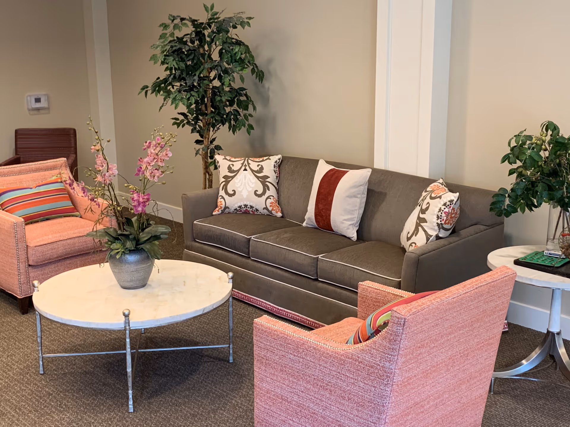 A cozy living room area with a gray sofa adorned with decorative pillows, two pink upholstered armchairs with striped cushions, a round white marble coffee table with a floral arrangement, and potted plants in the corners.