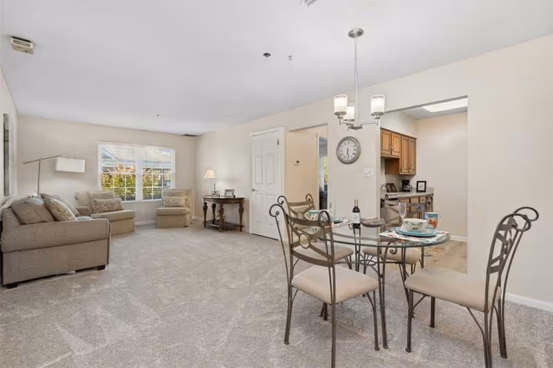 A bright and spacious living and dining area in a senior living facility. The living room features a beige sofa, two armchairs, a floor lamp, and a side table with a lamp and picture frames. The dining area has a glass-top table with four metal chairs, set with plates, bowls, and a bottle of wine. The kitchen is visible through an open doorway, showing wooden cabinets and stainless steel appliances. The room has light-colored walls and carpeted flooring, with a window letting in natural light.