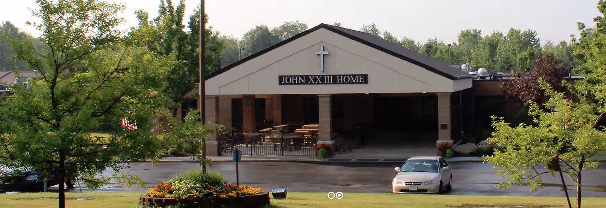 Front exterior view of the Saint John XXIII Home building with a peaked roof and a cross above the entrance. There is a covered porch area with tables and chairs, a parking lot with a car in front, and trees and greenery surrounding the building.