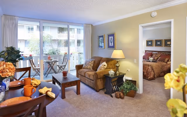 A cozy living room with a brown loveseat, wooden coffee table, and a side table with a lamp. The room has beige walls and carpeted floor. There is a dining table with orange cups and plates in the foreground. Sliding glass doors lead to a balcony with a small table and chairs. A bedroom with a bed and framed pictures is visible through an open doorway.