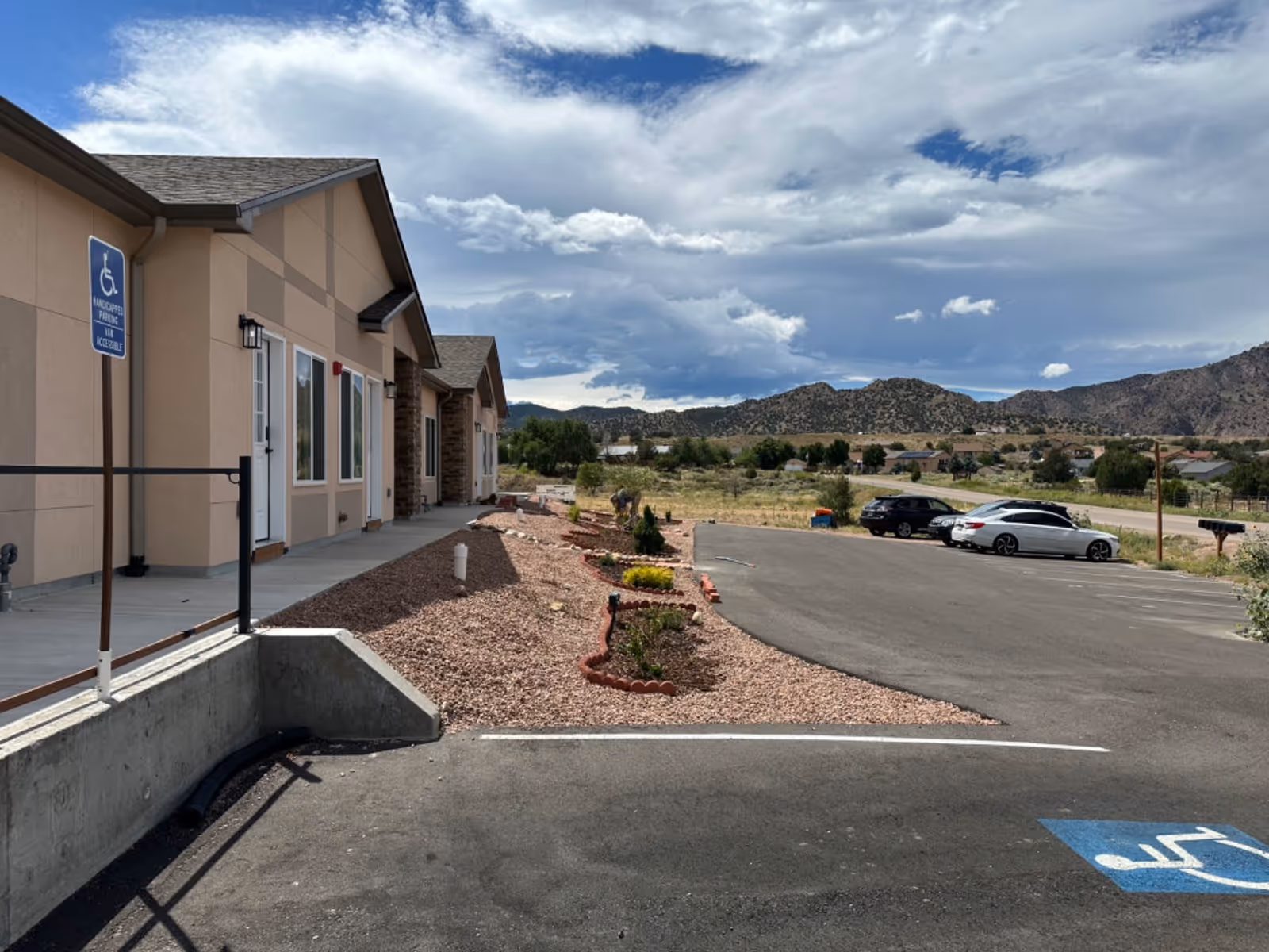 Exterior view of a single-story building with beige walls and a sloped roof, featuring a concrete walkway and a handicapped parking space in the foreground. There are several parked cars and a landscaped area with small plants and red mulch. In the background, there are hills under a partly cloudy sky.