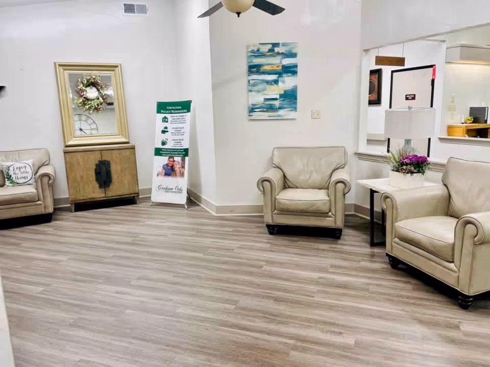 A waiting or sitting area with beige leather armchairs and a loveseat arranged around a small white side table with a lamp and a flower pot. The room has light wood flooring, white walls, a ceiling fan, a large mirror with a decorative wreath, and a standing sign with visitor policy reminders. There is also a blue and white abstract painting on the wall.