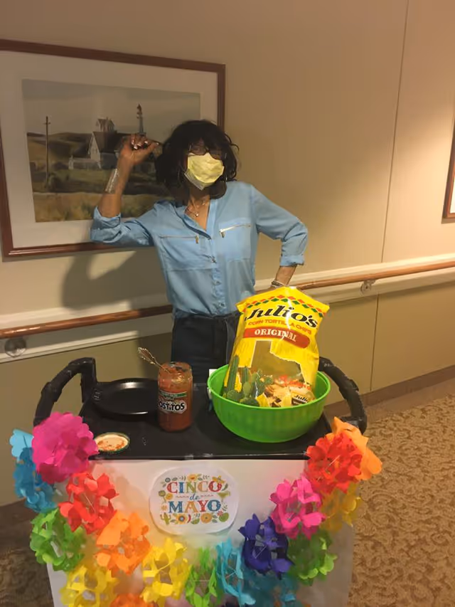 A woman wearing a yellow face mask and blue shirt stands behind a decorated cart in a hallway. The cart is adorned with colorful paper flowers and a sign that reads 'Cinco de Mayo.' On the cart, there is a large green bowl filled with bags of Julio's corn tortilla chips and a jar of Tostitos salsa with a spoon inside. A framed landscape painting hangs on the wall behind her.