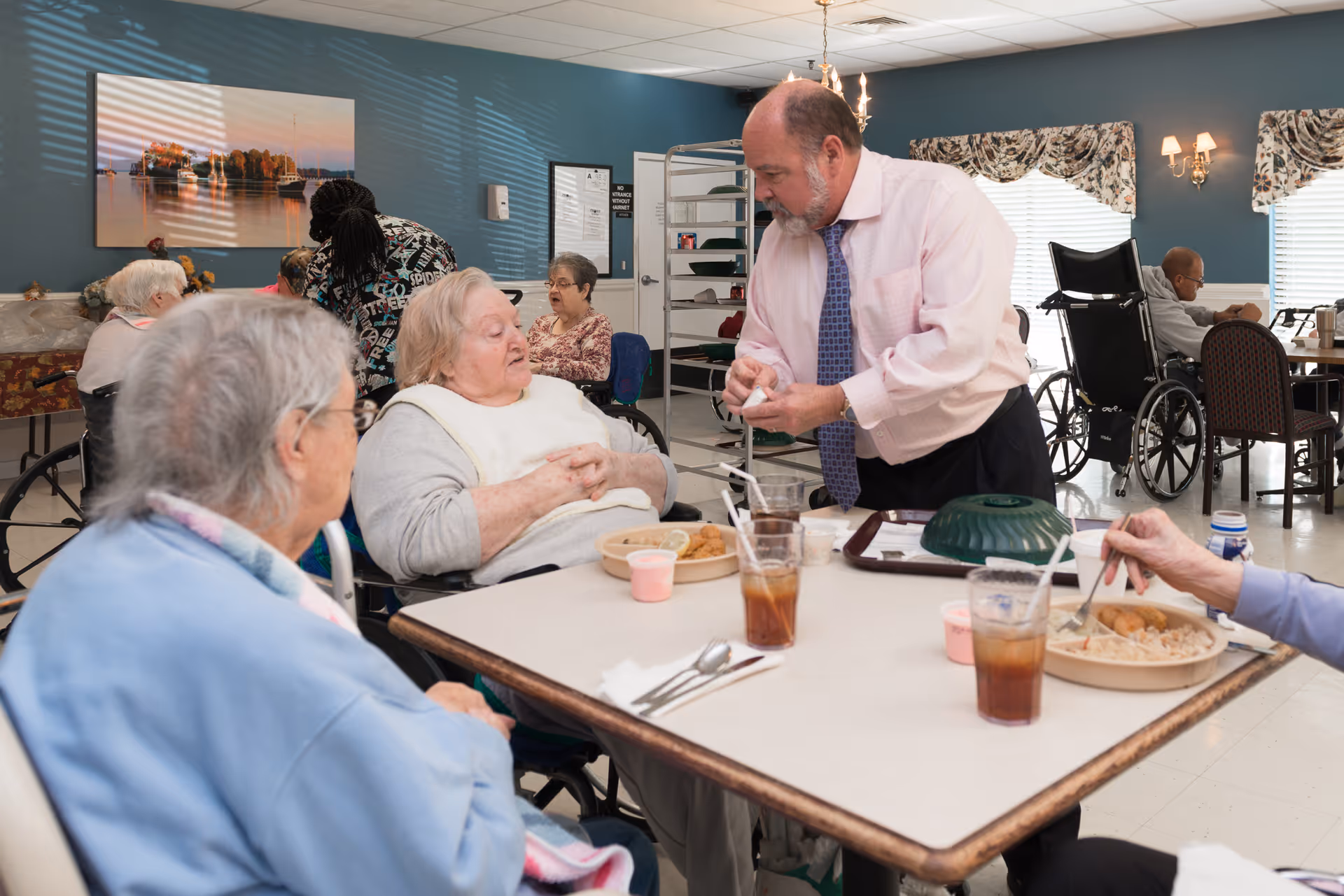 A man in a pink shirt and tie is assisting elderly women seated at a table in a dining area. The women are eating meals and drinks are on the table. Other elderly individuals are visible in the background, some in wheelchairs, in a well-lit room with blue walls and windows with floral curtains.