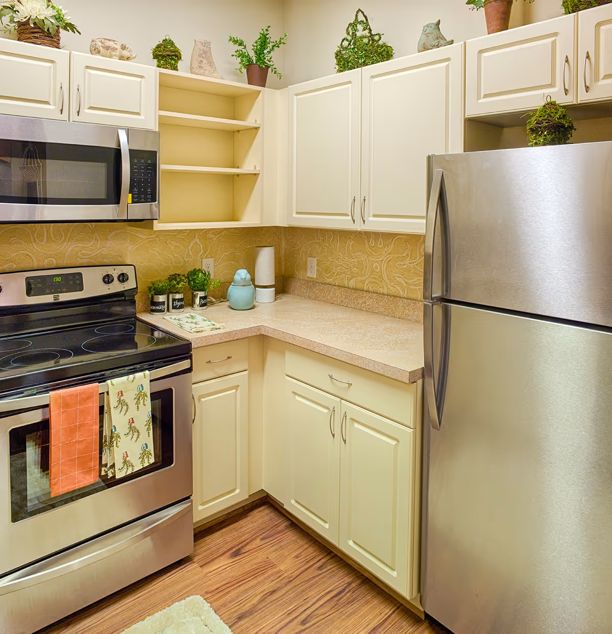 A clean and well-lit kitchen corner featuring stainless steel appliances including a microwave, electric stove, and refrigerator. The kitchen has cream-colored cabinets, a beige countertop, and decorative plants and ceramic items on top of the cabinets. There are two kitchen towels hanging on the oven handle, and the floor is wooden with a small rug partially visible.